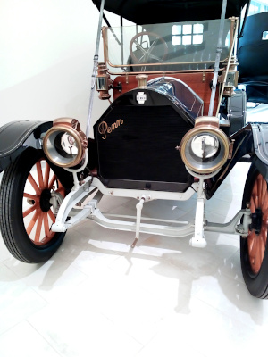 [There appears to be a hand crank at the front. There are two brass-rimmed headlight. The engine has a black front and the scripted word Penn in brass along one side. The windshield covers the lower half of the front opening of the vehicle and it is likely the driver would not be protected much by it because it barely seems to clear the steering wheel.]